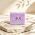 Jabón artesanal Mayam Holistic de lavanda y manzanilla en tono lila sobre base de piedra natural con sombras de hojas.