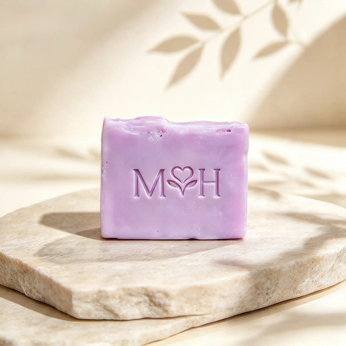 Jabón artesanal Mayam Holistic de lavanda y manzanilla en tono lila sobre base de piedra natural con sombras de hojas.